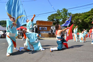夏の青空の下、お揃いの水色の着物を着た踊り手たちが鮮やかな旗を振りながら元気よくポーズを決めているよさこい踊りの写真