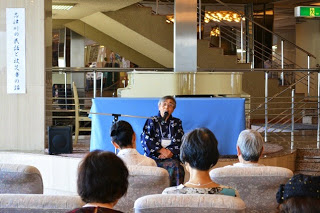 階段横の前方に設置されたマイクスタンドの前に座った、青色のシャツを着た人物の話しを参加者が聞いている後ろ姿の写真