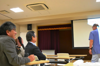 男性の参加者が前方を見ながらマイクを使って講師に質問をしている様子で、講師はスクリーンの方を向いている写真