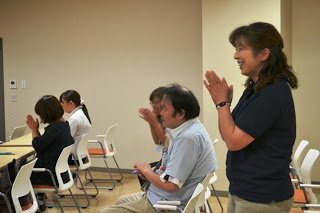 椅子に座ったり立ち上がって拍手をしている笑顔の参加者たちを横から撮影した写真