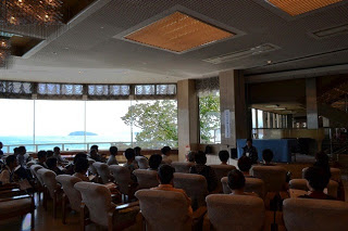 大きな窓から海が見える開放的な空間で、参加者たちが静かに一人の話者の方へ向かって座っている写真