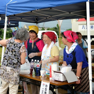 屋台のテントの下でカラフルな三角巾をかぶった女性たちが、笑顔でお客さんにかき氷を販売している写真