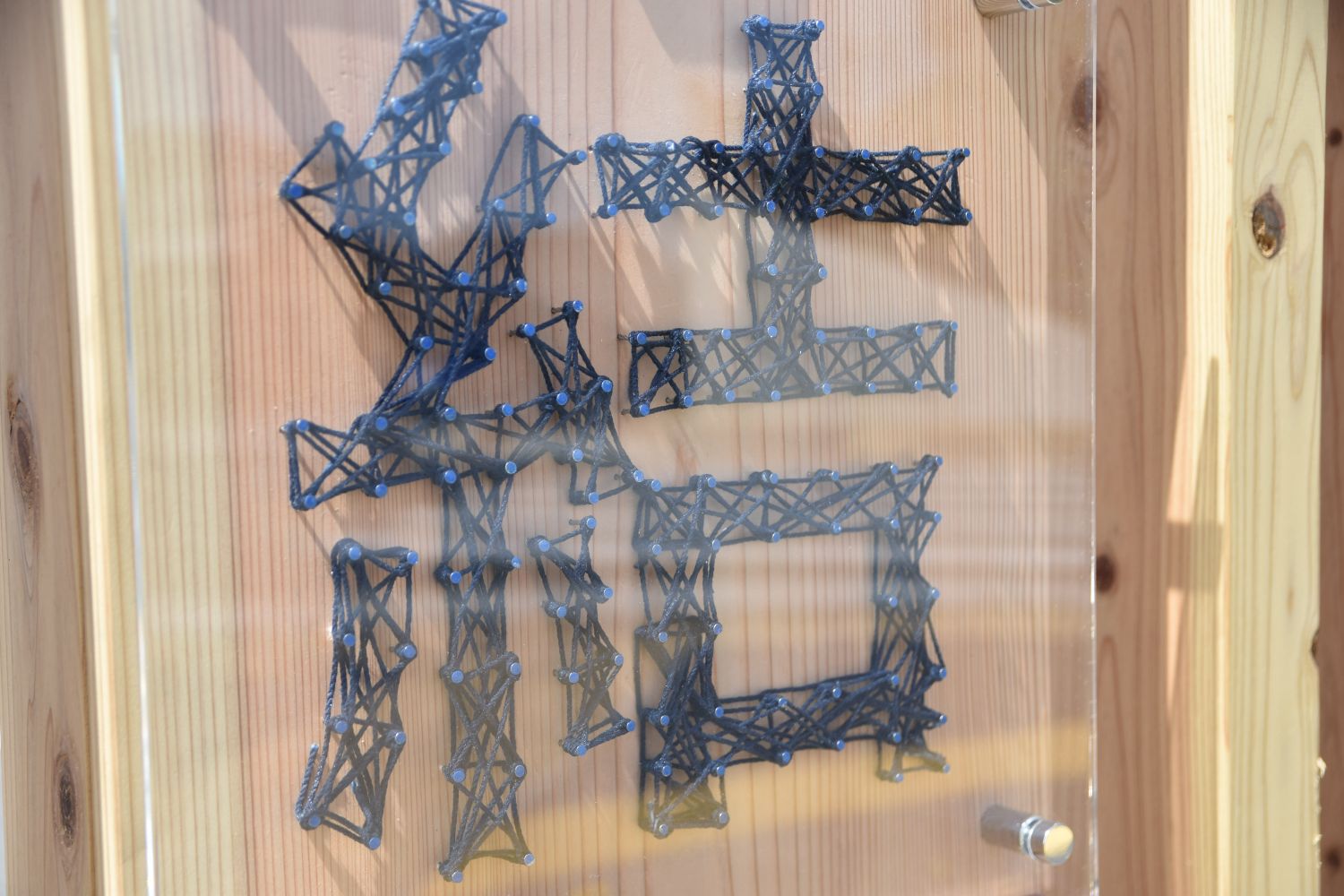 釘と糸で作られた「結」の文字が木の板に装飾されている看板を、透明なカバー越しに撮影した写真