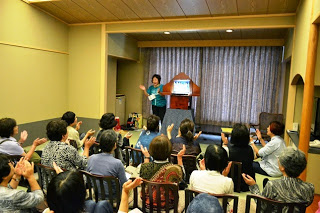 和室風の会場で、舞台に立つ女性が紙芝居を披露し、観客たちが拍手をしながら楽しんでいる写真