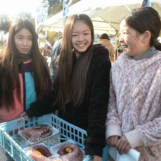 三人の少女がパック詰めされたタコの商品が入った青いカゴを持って、笑顔で写っている写真