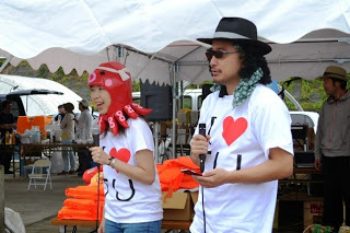 タコの帽子をかぶった女性とサングラス姿の男性がマイクを持って司会をしているステージ前の写真