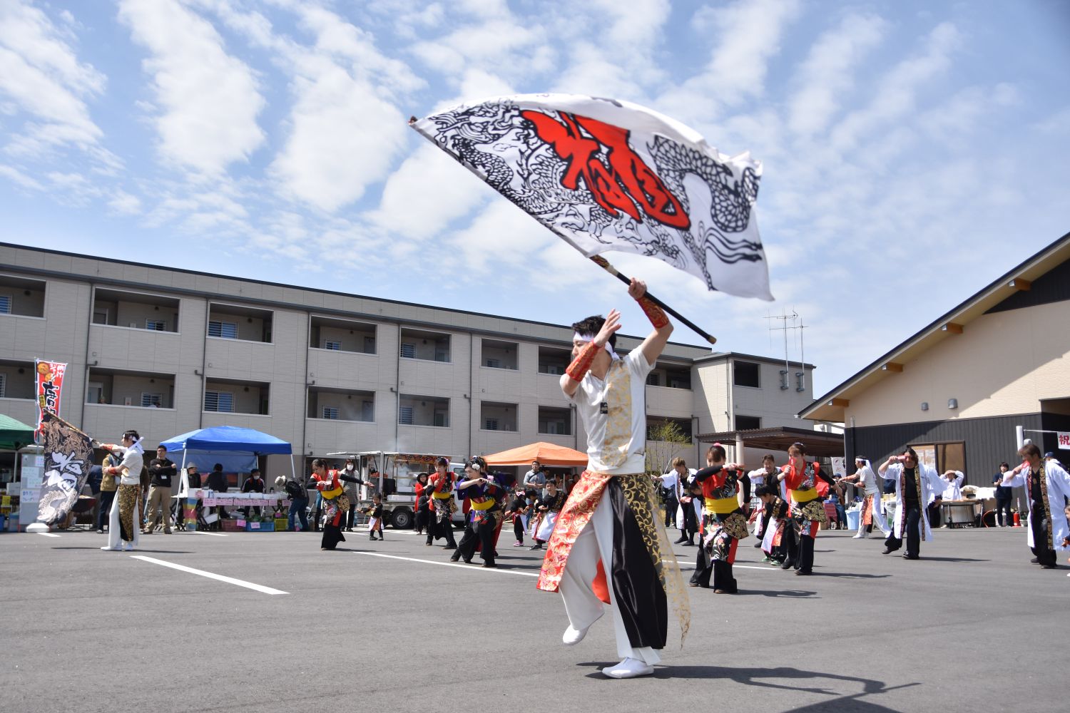 大きな旗を振る男性を先頭に、色とりどりの衣装を着た踊り手たちが屋外の広場でパフォーマンスをしている写真