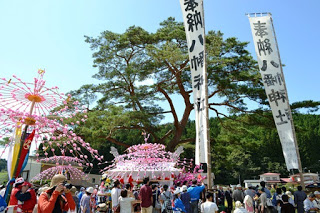 大きな松の木の前に、ピンクの紙飾りで華やかに飾られた山車（だし）と、それを見に集まった多くの人々の様子が写っており、「奉納八幡神社」と書かれた白い旗が立っている写真
