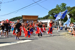赤と黒の衣装を着た若者たちが広場いっぱいに広がって手足を伸ばしながら踊っており、観客がその活気ある踊りを見ているよさこいチームの演舞の写真
