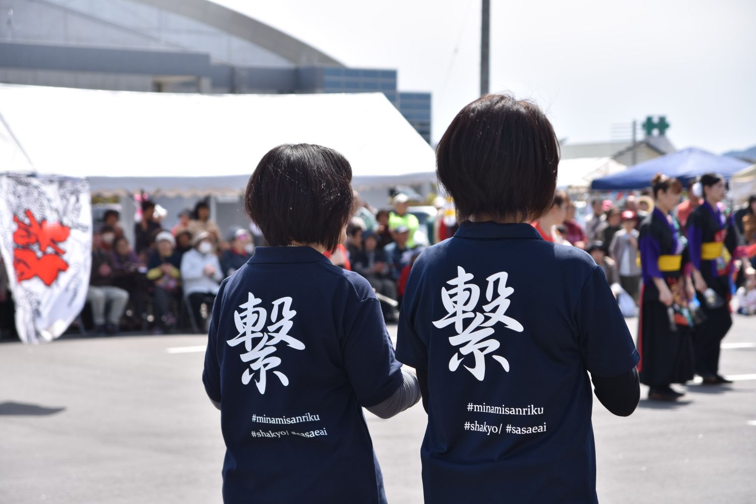 背中に「繋」と書かれた紺色のTシャツを着た2人の女性が、にぎわうイベント会場に立っている様子の写真