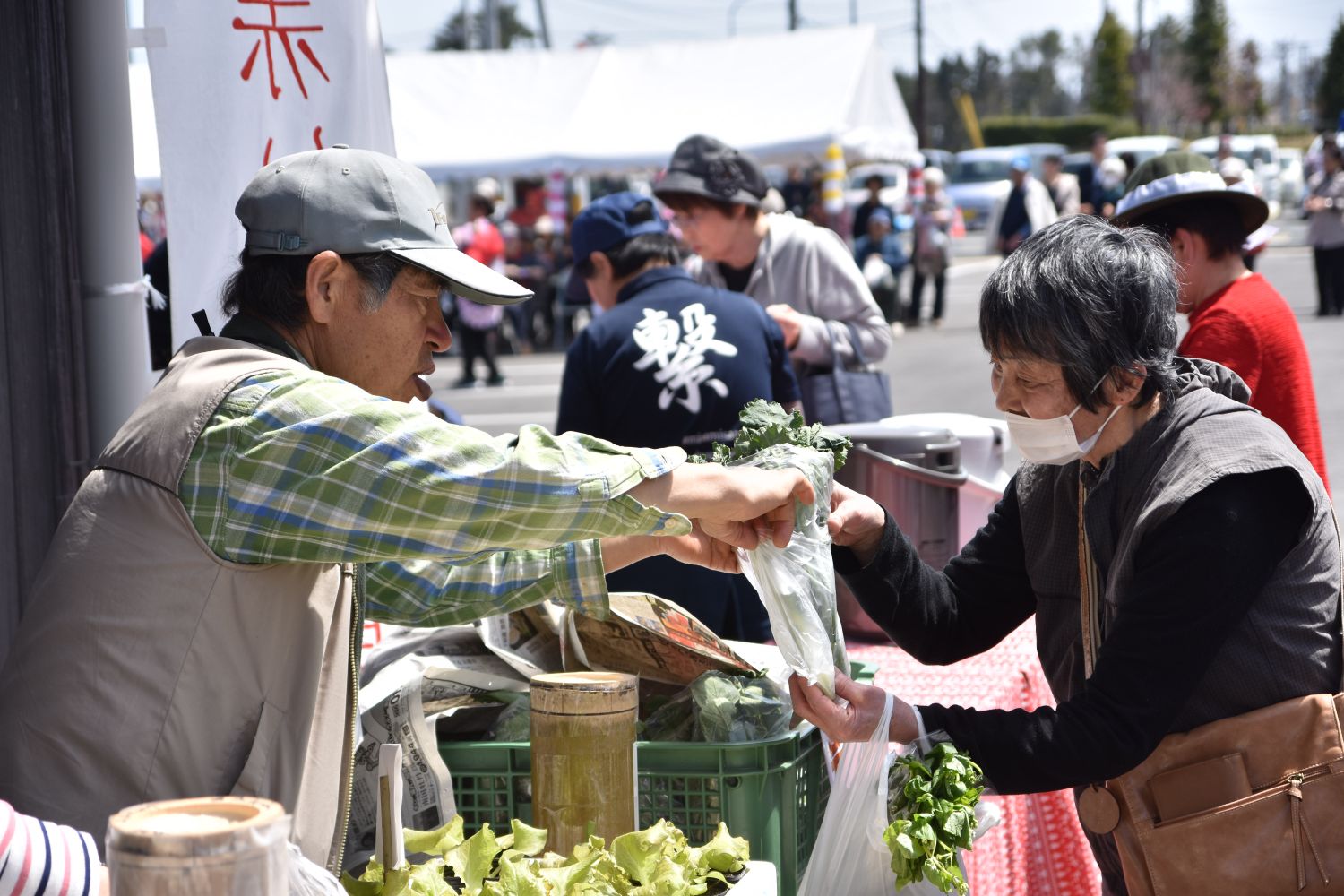 イベント会場のテントの下で、男性が新鮮な野菜を袋に詰めて女性客に手渡している写真