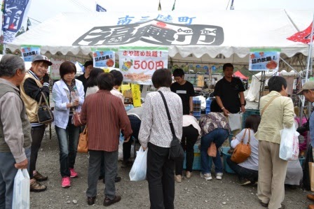 「ホヤ詰め放題！500円」と書かれた看板のあるテントの前に、大勢の人が並んで商品を見たり買ったりしている写真