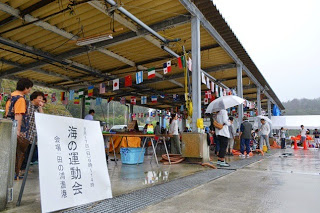 屋根付きのスペースに国旗の飾りが吊るされ、多くの人が集まって準備や見物をしている様子で、手前に「海の運動会」と書かれた看板が立っている写真