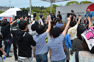 屋外の特設ステージで音楽ライブが行われており、前方の観客たちは腕を上げて盛り上がり、バンドメンバーが演奏している様子の写真