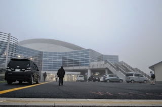 曲線的な屋根と大きなガラス窓を持つ現代的な建物が霧の中に立ち、前方には数台の車が停まり、数人の人が建物に向かって歩いていて、右側に2階の入口へ続く外階段が見える写真