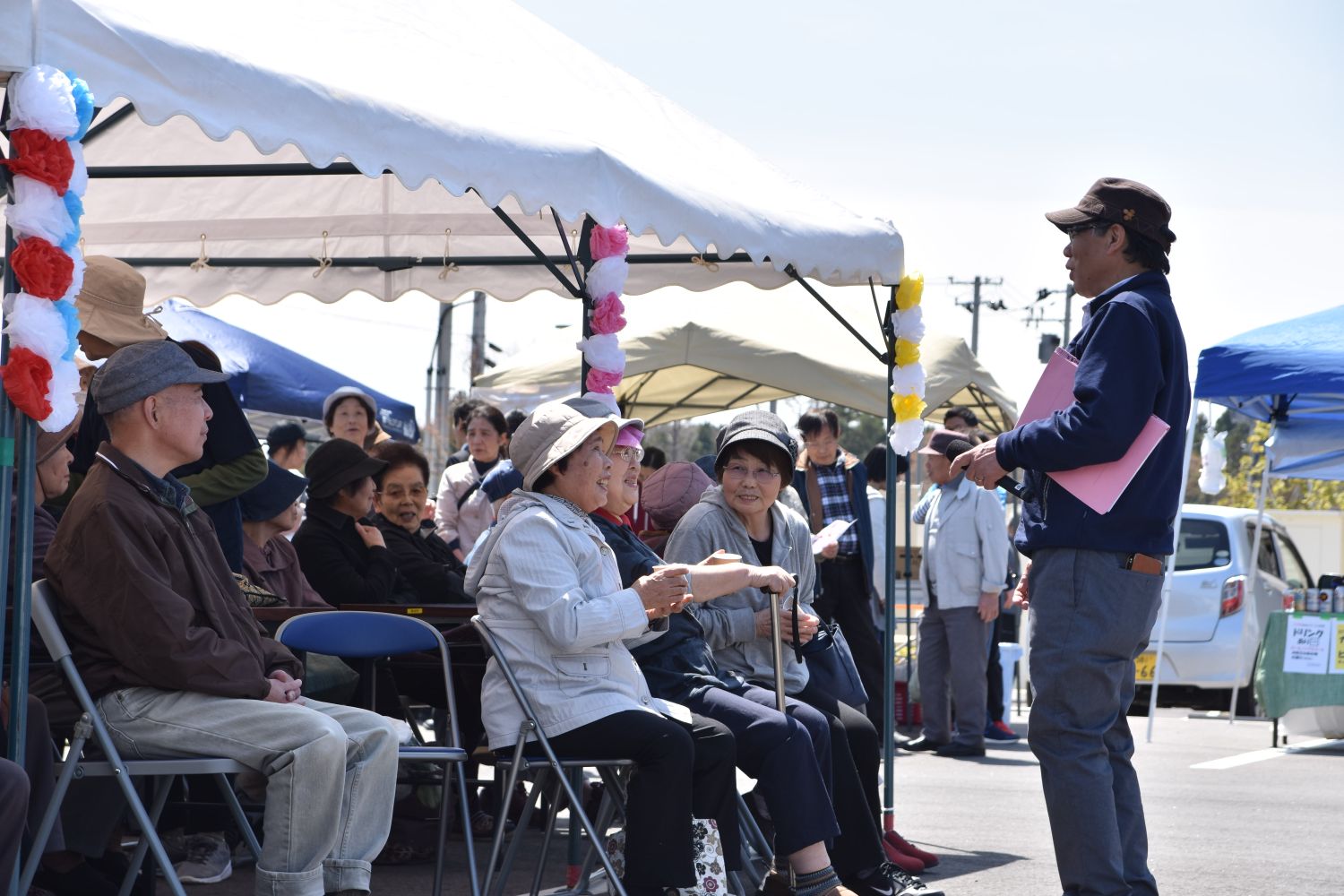 テントの下に座った高齢者たちが談笑しながら、ピンクのファイルを持った男性の話に耳を傾けている写真