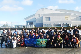 大きな白い壁の建物を背景に、大勢の人々が整列し、前列中央の人たちが横断幕を持ち、全員でカメラに笑顔を向けている集合写真