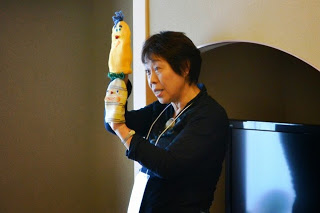 女性がカラフルな人形を両手に持ち、観客に向かって人形劇を披露している写真