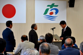 白い壁に国旗と町旗が掲げられた室内で、2人の男性と向かい合い、マイクスタンドの前に立つ町長が感謝状を読み上げている様子の写真