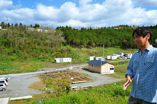 花壇や木造建物、木々に囲まれた森を背景に、丘の上でチェック柄のシャツを着た男性が話しをしている写真