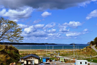 緩やかな坂の先に海が広がり、空には雲が多く浮かび、手前には復興施設が並んでいる写真
