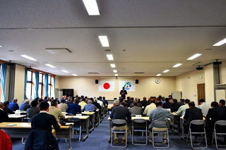 壁に国旗と町旗が掲げられた室内で、大勢の参加者たちが席に座り、前方に立つ男性の話を聞いている様子を後ろから撮影した写真