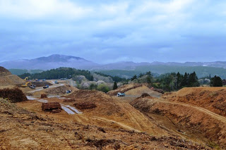 奥には山並みが見える曇り空の下、大な造成地に重機や車両が点在している、造成工事が進む山間部の全景写真