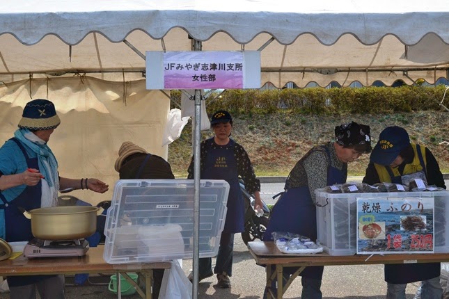 柱に「JFみやぎ志津川支所女性部」と書かれた看板が掲げられているテントの下で、女性スタッフがカセットコンロと大鍋の後ろで準備を行い、沢山の乾燥ふのりが入っている大きな衣装ケースの後ろで販売の準備をしている様子の写真
