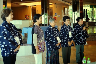 ロビーのような場所で、お揃いの紺色のシャツを着た女性たちが並び、右端に立つ女性がマイクを持ち話をしている写真