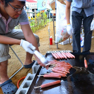 屋外で鉄板の上に並べたフランクフルトを男性がトングで焼いている様子の写真