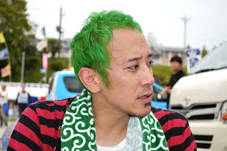 赤と黒のボーダー柄の服を着て、緑と白の唐草模様のタオルを首にかけて横を向いた、鮮やかな緑色の髪をしたGo！さんの写真