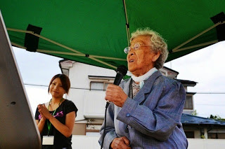 緑のテントの下で、手拍子をした女性が見守っているなか、マイクを持った高齢の女性が歌っている様子の写真