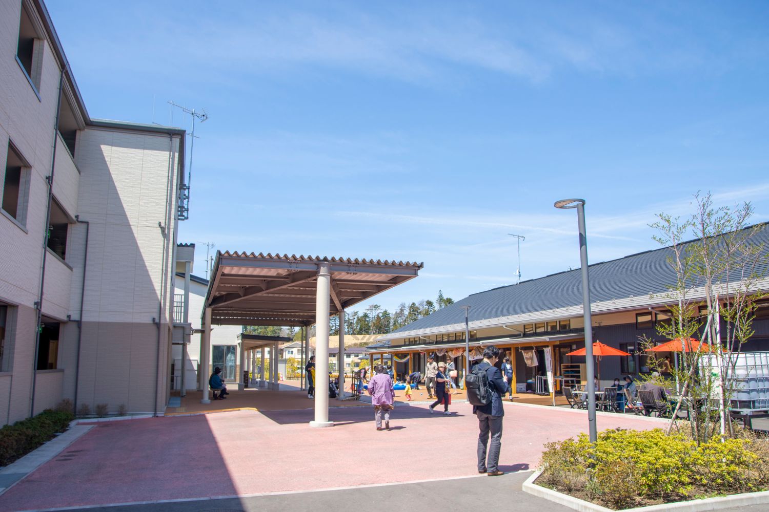 左に災害公営住宅、右に屋根付きの平屋建ての「結の里」があり、間の広場を数人の人が歩いている晴れた日の住宅地の写真