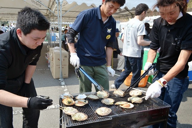 屋外イベントの会場に設置されたバーベキューコンロで、両手に軍手を着けた3人の男性がトングを使って貝類を焼いている写真