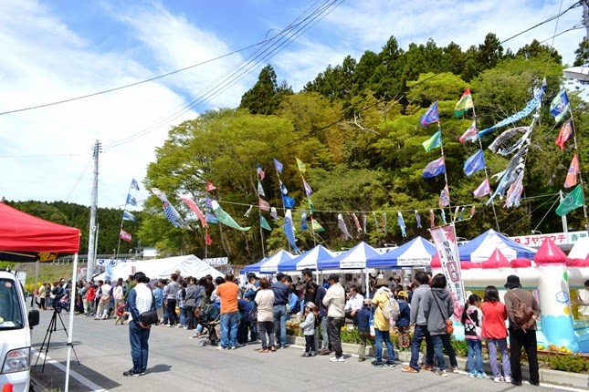 大漁旗が風に揺れている木々に囲まれた通路沿いで、大勢の人がずらりと列をなして並んでいる写真
