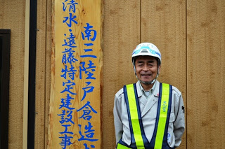 青色で文字が書かれた木板の前で、安全ヘルメットを被り、作業服の上から反射ベストを着た所長が笑顔で立っている様子をアップで撮影した写真