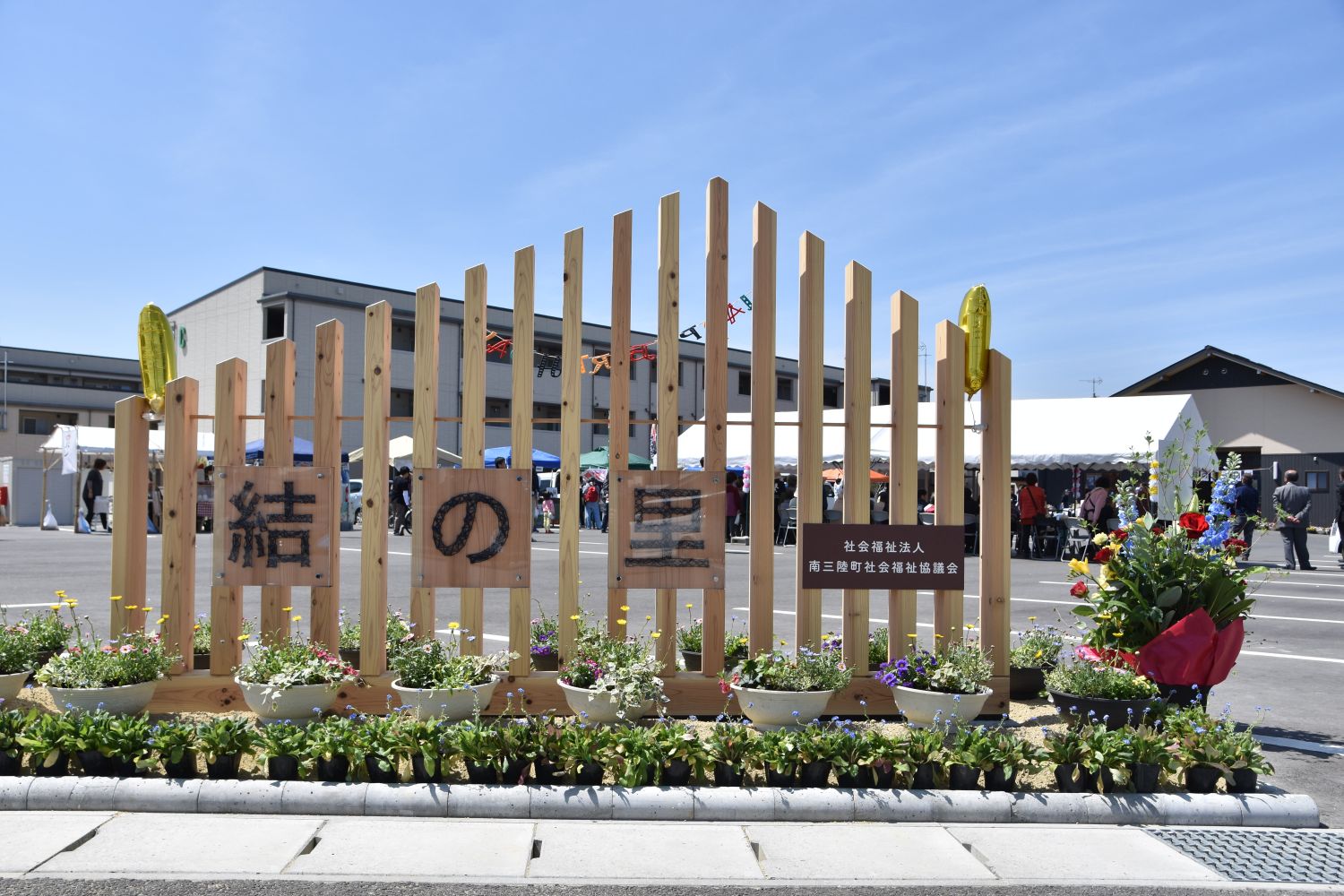 「結の里」と書かれた木製の柵が中央にあり、手前には色とりどりの花が並べられているイベント会場の入口の写真