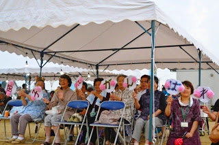 白いテントの下で椅子に座った女性たちが、手作りのうちわを持って笑顔で声援を送っている写真