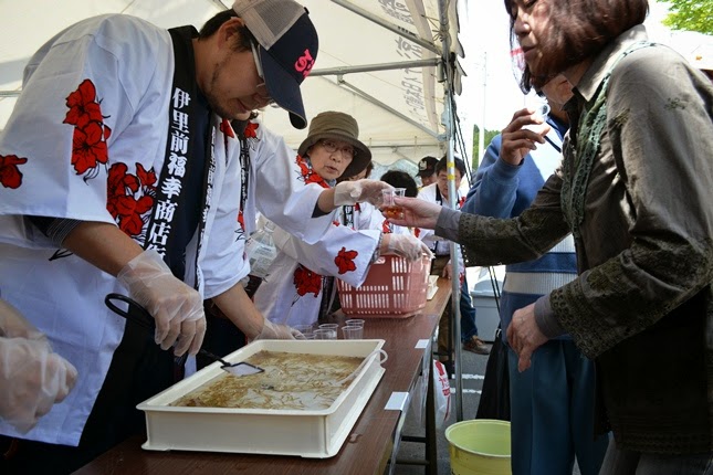 法被を着たスタッフの男性が白いテントの下で、ネットが貼られたぽいを右手で持ち、お客さんに酢醤油の中のしらうおが入った小さな透明カップを配っている写真
