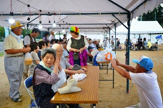 屋外のテントの下で高齢者の方々が椅子に座って談笑している写真