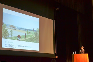 「ポートパーク」と書かれた説明と公園のイメージが映し出されているスクリーンがステージに設置され、隈氏がプレゼンテーションを行っている様子の写真