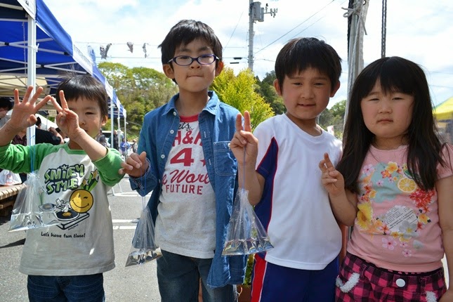 4人の子どもが笑顔でピースをしながら、手にしらうおの入ったビニール袋を持って並んでいる写真