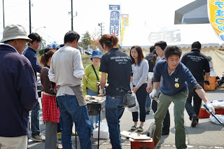屋外の祭り会場で、複数の人々がホタテを焼いているバーベキューコンロの周りに集まり、料理や食べ物の提供をしている様子が写されている写真