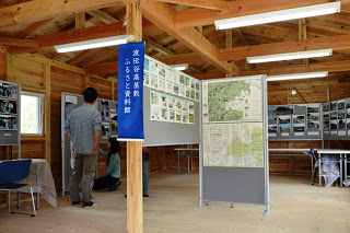 木造の資料館の中で、大人3人が壁や掲示ボードに貼られた地図や写真が展示されたパネルを見学している写真