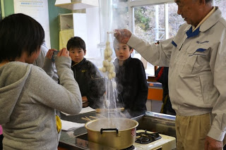 阿部先生が湯気の上がる鍋から箸で繭を引き上げ、白い繭からたくさんの絹糸がほつれて伸びている様子を子どもたちが見つめている、絹の糸取り体験の写真