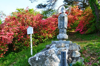 満開のピンクのツツジが咲く急斜面に、円形の光背がある岩の上に立つ観音像と説明板がある写真