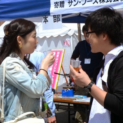 若い男女がイベント会場のテントの前でしらうおが入ったカップを手に楽しそうに会話している様子の写真