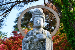 赤いツツジが広がっている背景に、両手を胸元に合わせ、静かにたたずむ灑水観音像の上半身をアップで撮影した写真