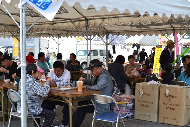 屋外イベントに設置されたテントの下の飲食スペースで、沢山の人々がテーブルに座って飲食をしている様子の写真