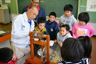 教室で阿部先生が絹糸を取る機械を操作し、複数の小学生が興味深く見学している様子の写真